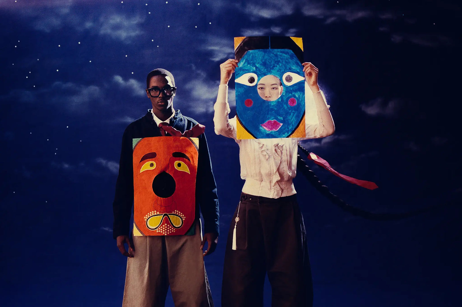 Two people standing against a dark starry night sky backdrop, each holding a large abstract painted face mask or collage. The person on the left wears glasses and a dark jacket with light pants, while the person on the right wears a white top and dark pants.
