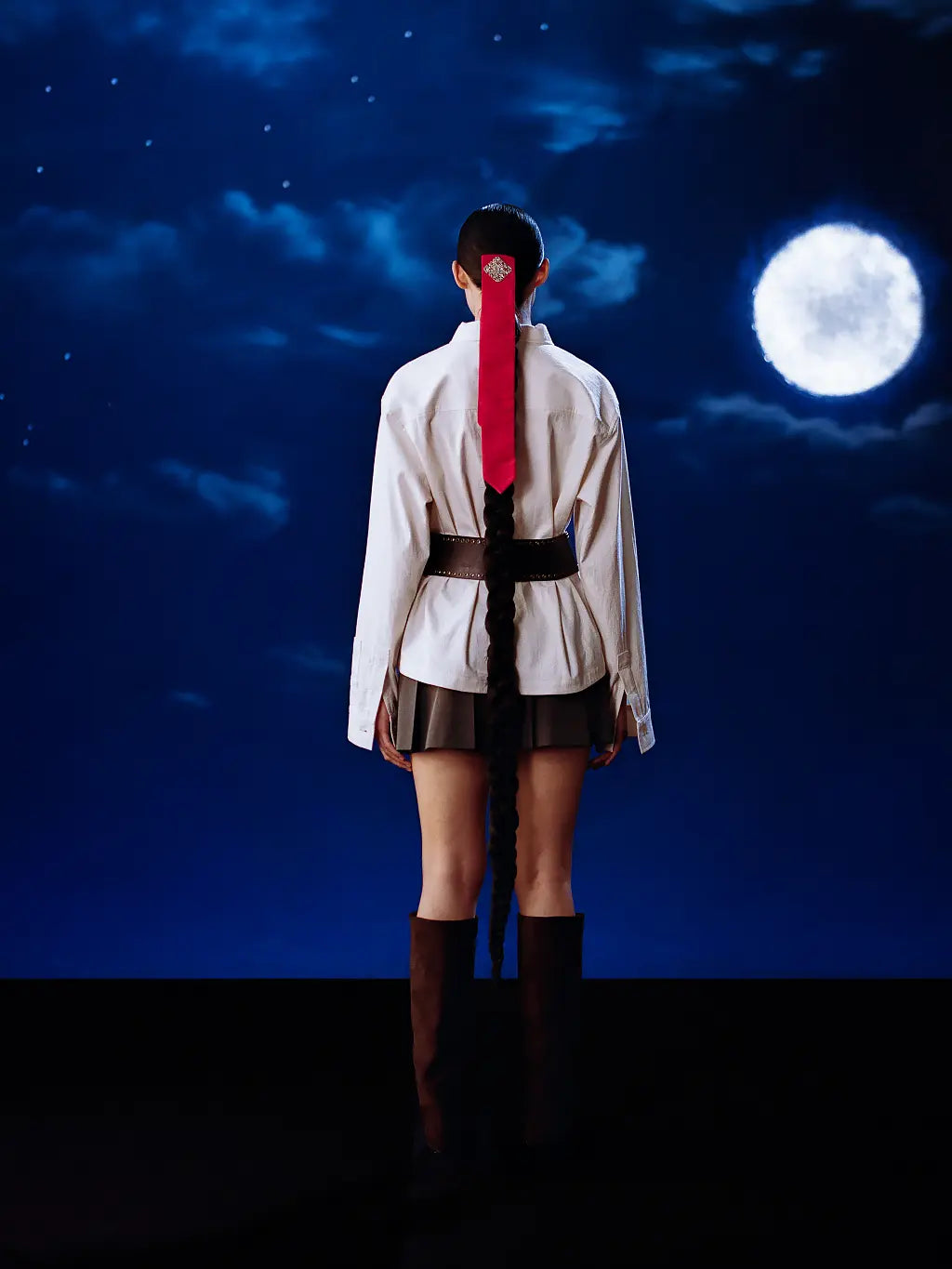 A model stands with their back to the camera wearing Sundae School Ivory China Collar Knot Shcaket, a dark pleated mini skirt, and dark brown knee-high boots. Their hair is styled in a long braid with a bright red ribbon accent.