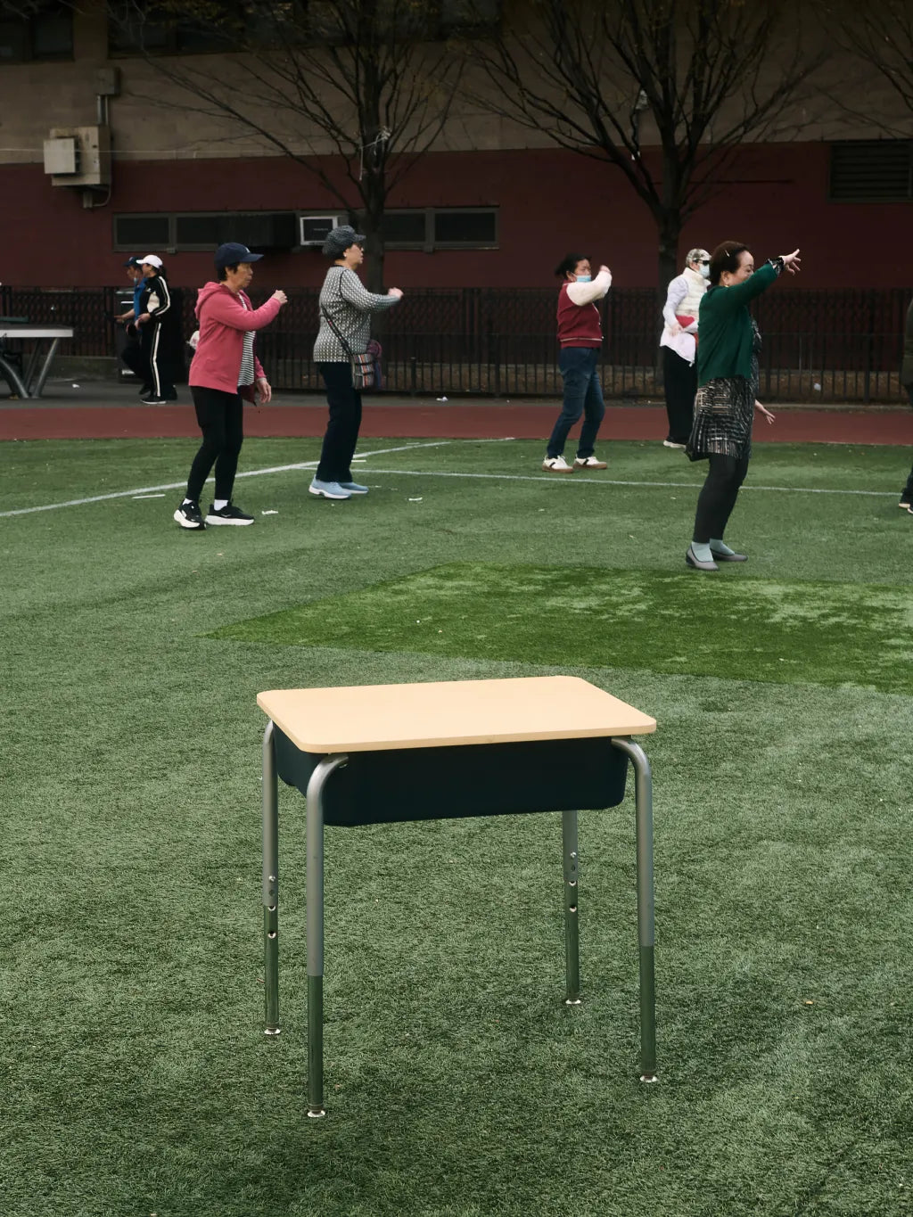 A school desk with a light wood top and dark green fabric cushion sits on green artificial turf in an outdoor playground or sports field, with several people exercising in the background.