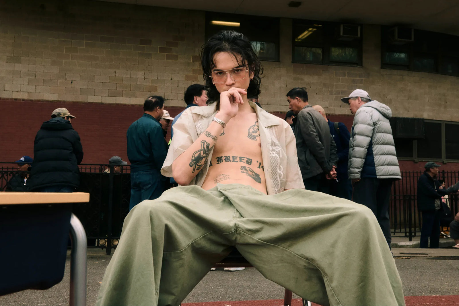 A person with dark hair and glasses sits on a chair outdoors, wearing BARONG SHIRT over a bare torso with visible tattoos, and OLIVE HANBOK CHINO PANTS. Several people are standing in the background near a brick building.