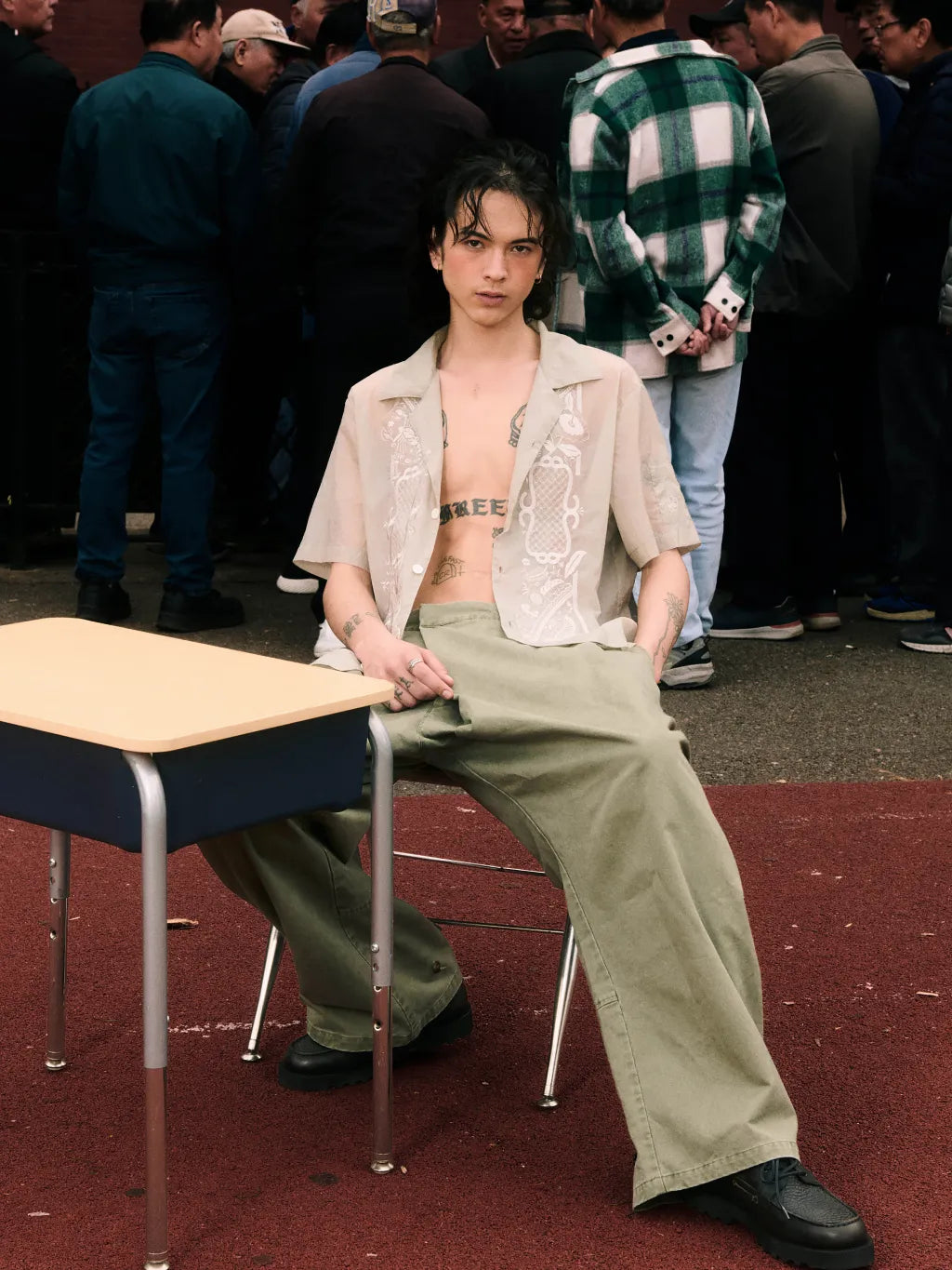 A person seated on a chair in front of a school desk, wearing SUNDAE SCHOOL BARONG SHIRT over a bare torso with SUNDAE SCHOOL OLIVE HANBOK CHINO PANTS and black shoes. A crowd is visible in the background, and the scene appears to be an outdoor or event-like setting.