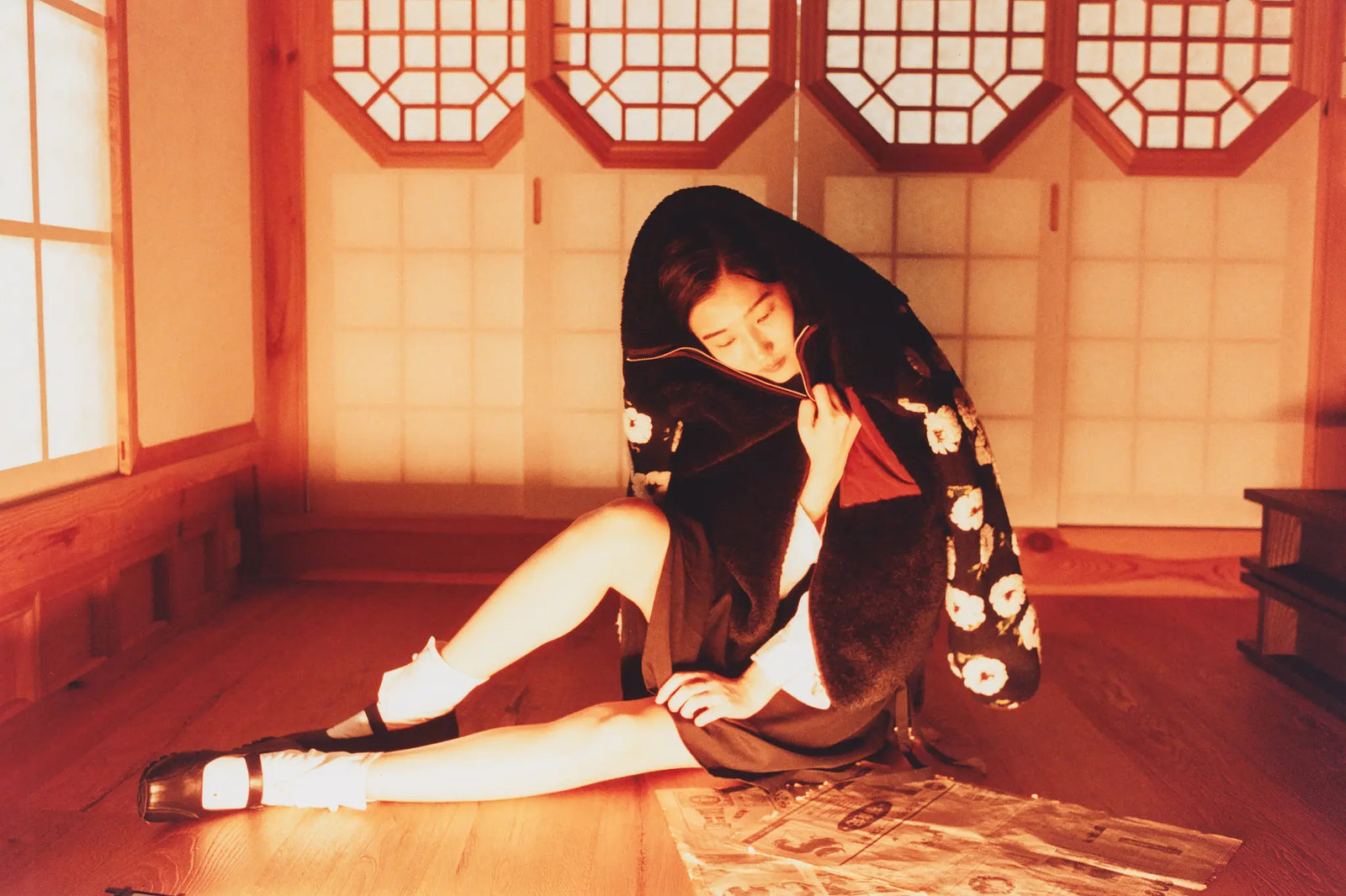 A woman in Sundae School Mugunghwa Fleece with white floral patterns sits on the floor in a warm, orange-lit room with wooden walls and sliding paper doors.
