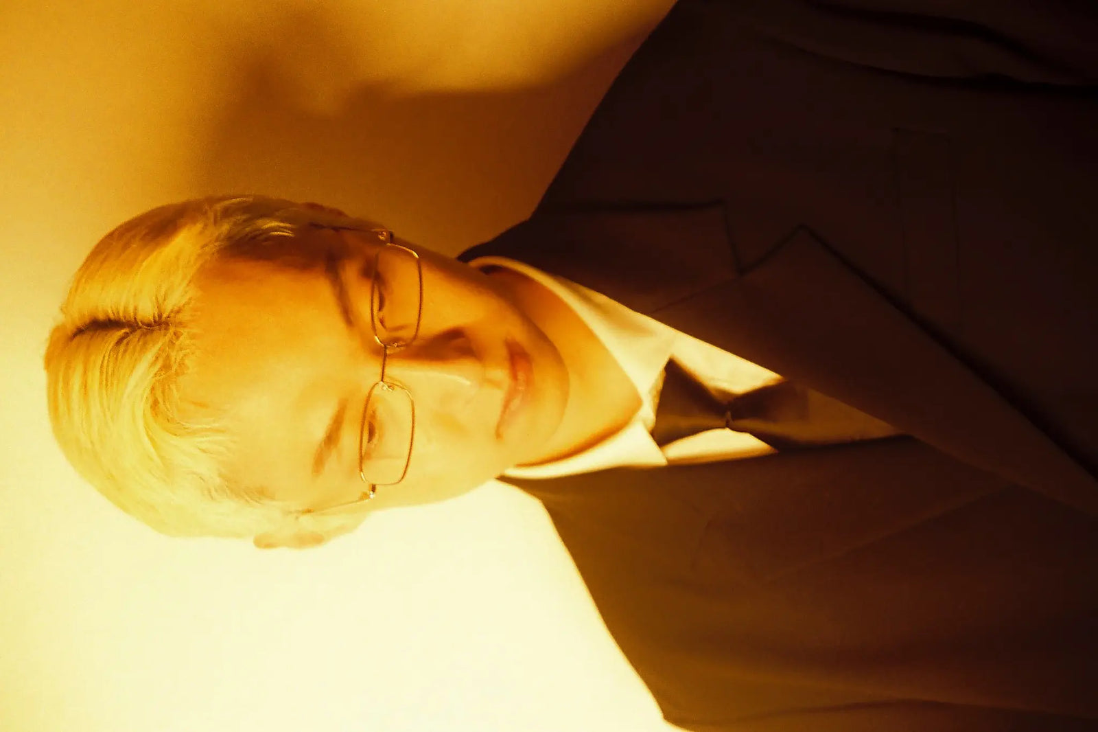 A close-up portrait of a man wearing glasses, a white collared shirt, and a dark suit jacket, photographed in warm yellow lighting against a matching warm-toned background.