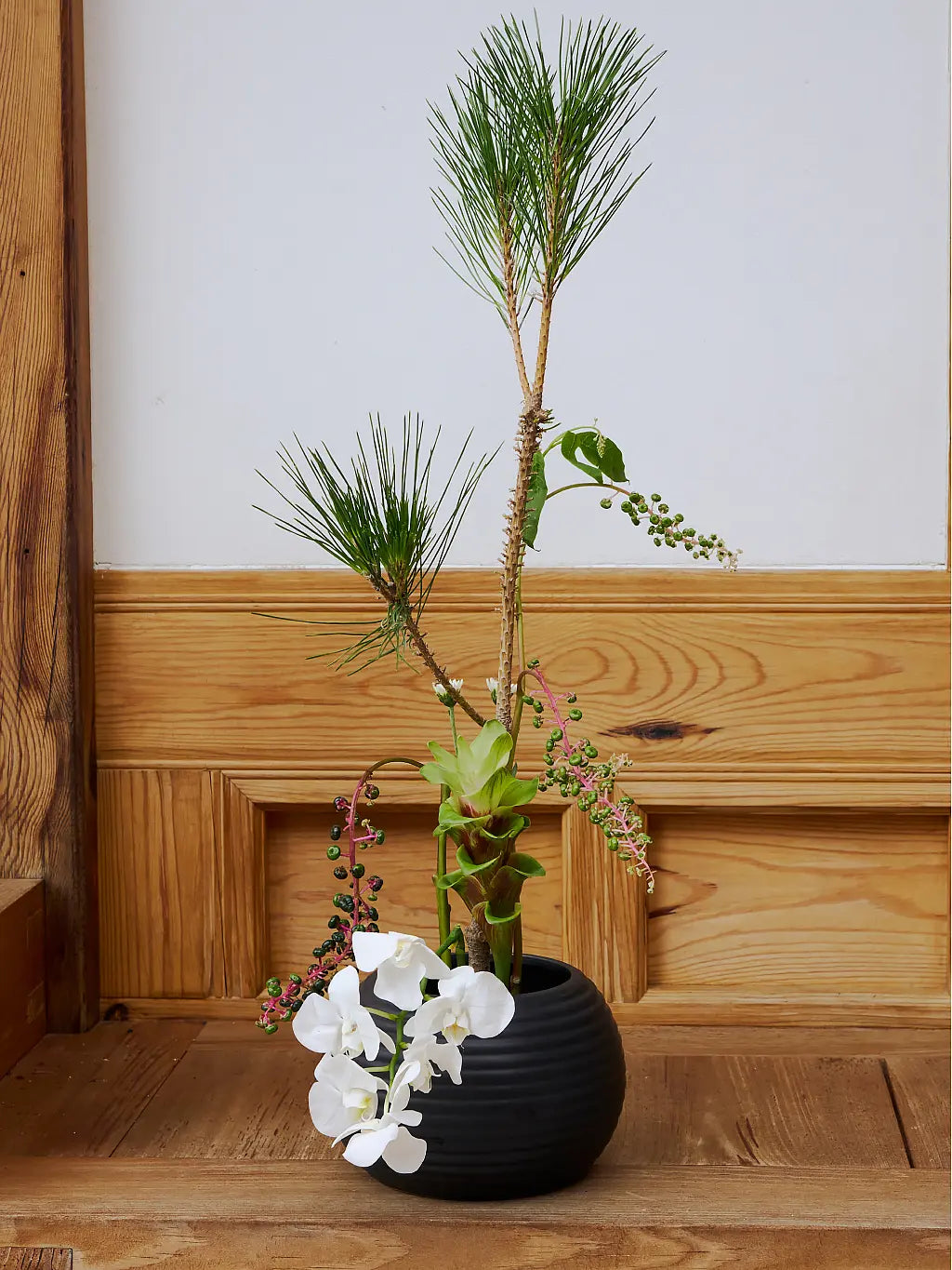 A black ribbed ceramic planter holding a minimalist Japanese-style floral arrangement with a tall pine branch, green foliage, small pink blossoms, and white flowers, set on a wooden surface against a white wall with wood paneling.