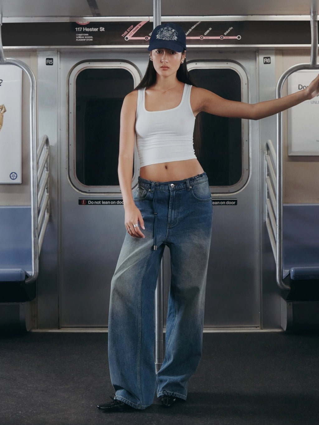 A woman stands on a subway train wearing a white ribbed tank top, wide-leg blue jeans, and a navy baseball cap.