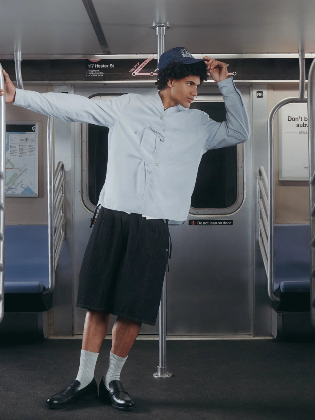 A model posing on a subway train wearing a light blue long-sleeve shirt, a black pleated skirt, white socks, black loafers, and a navy blue baseball cap.