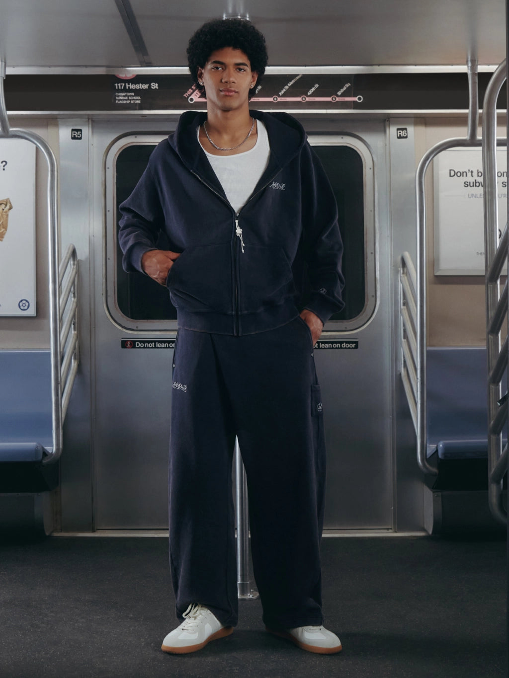 A man wearing a navy blue zip-up hoodie and matching sweatpants stands inside a subway train car, with silver doors and blue seats visible in the background.