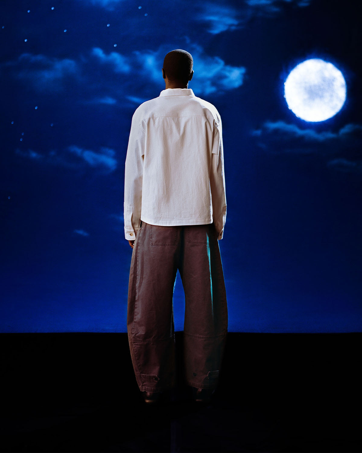 A person in a white shirt and Sundae School CHARCOAL HANBOK CHINO PANTS stands silhouetted from behind against a glowing full moon on a dark, cloudy night.