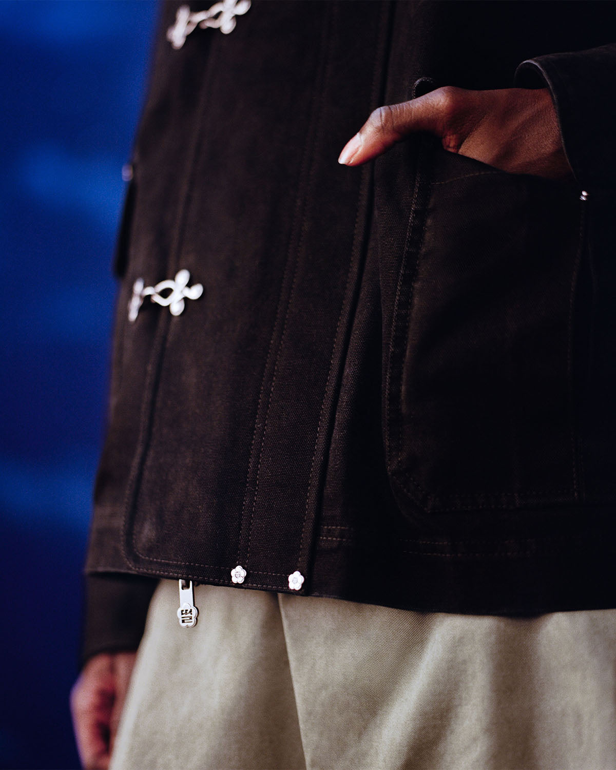 A person wearing the Sundae School [PRE-ORDER] BLACK FIREMAN CLASP JACKET with silver clasps and light pants stands with one hand in a pocket against a blurred blue background.