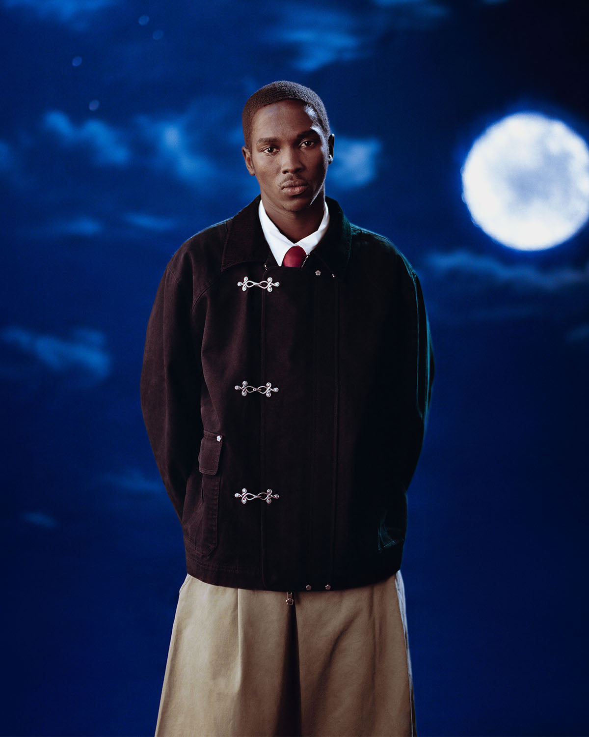 A man gazes at the camera under a full moon, wearing the Sundae School [PRE-ORDER] BLACK FIREMAN CLASP JACKET over a white shirt and red tie, paired with light pants against the nighttime sky.