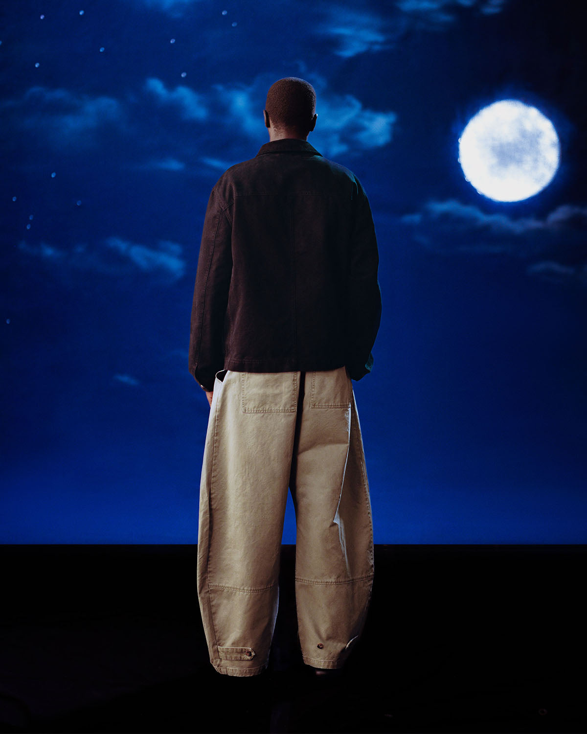 A person stands with their back to the camera, wearing the Sundae School [PRE-ORDER] BLACK FIREMAN CLASP JACKET and oversized light-colored pants, gazing at a night sky filled with clouds and a bright full moon.