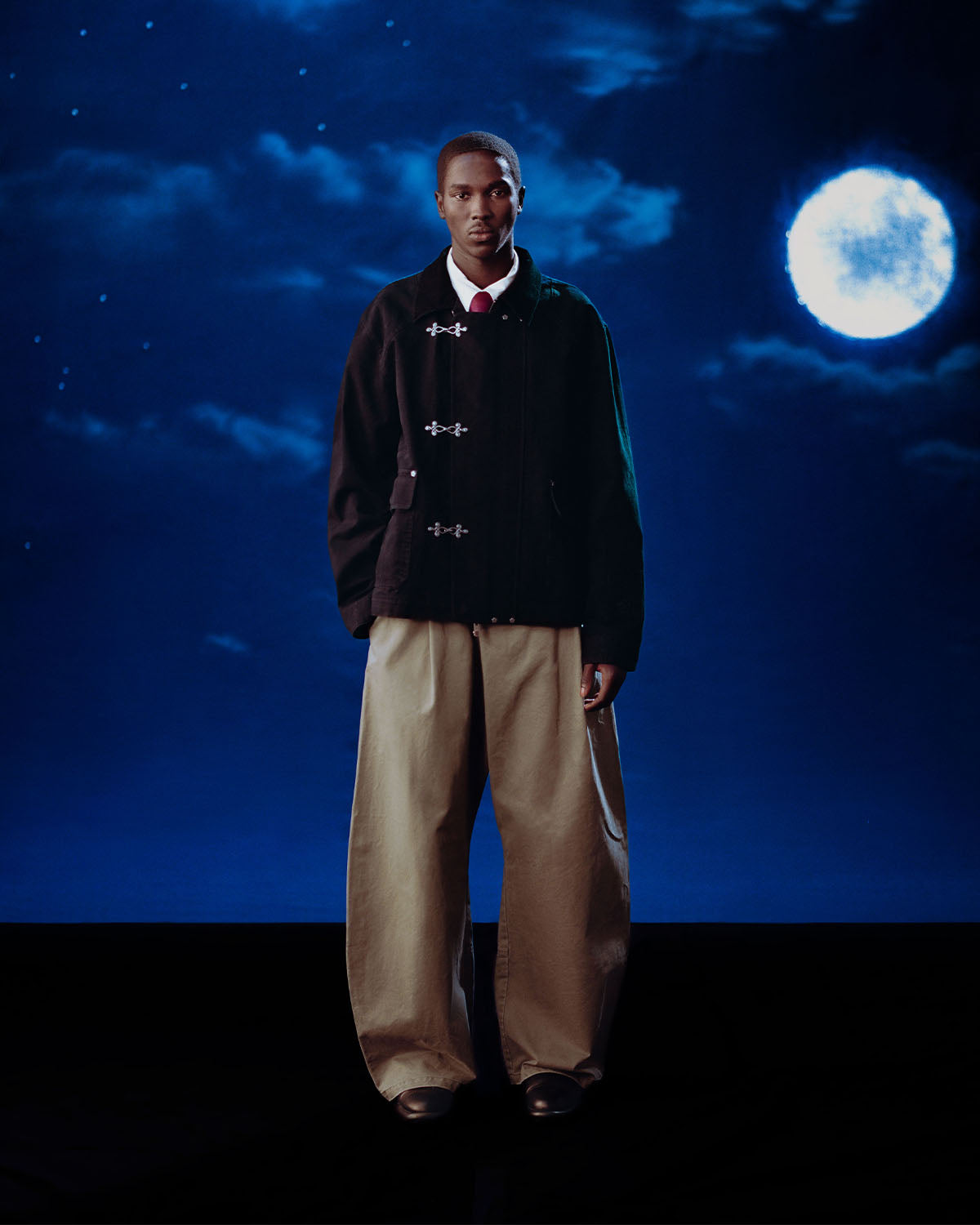 Beneath a full moon, a person faces forward with a neutral expression, wearing Sundae School’s [PRE-ORDER] BLACK FIREMAN CLASP JACKET over a white shirt and wide tan pants.