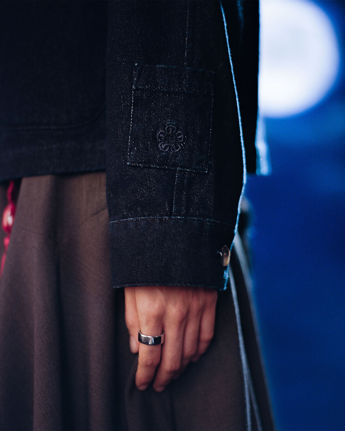 A close-up shows a hand wearing a silver ring, with an arm in the Sundae School DENIM CHORE JACKET featuring floral-stitched cuffs, paired with a brown pleated skirt. The blurred background is blue and white.