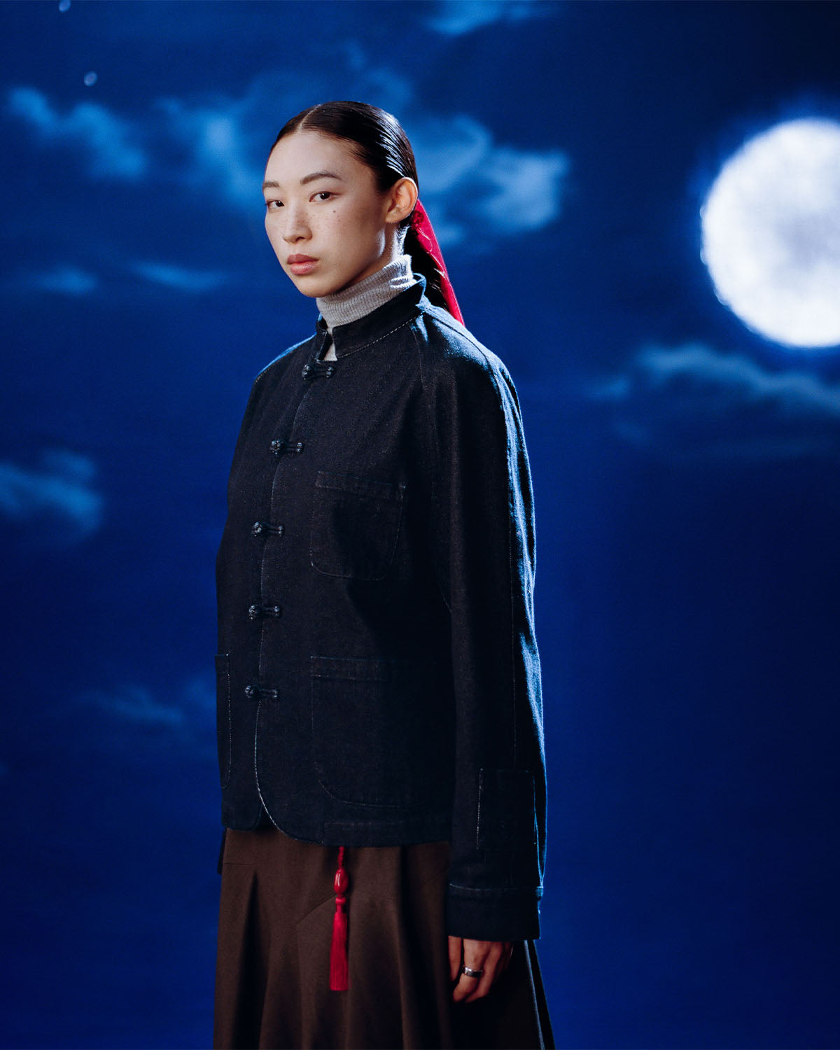 A woman with a red ribbon in her hair stands under a full moon and scattered clouds, wearing the Sundae School DENIM CHORE JACKET and a brown skirt, gazing calmly at the camera against the dark blue night sky.