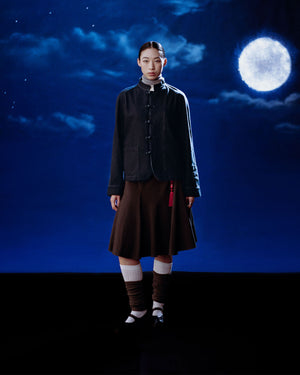 A young woman stands against a night sky backdrop with clouds and a bright full moon, wearing Sundae School’s DENIM CHORE JACKET, paired with a brown skirt, knee-high socks, and black shoes.