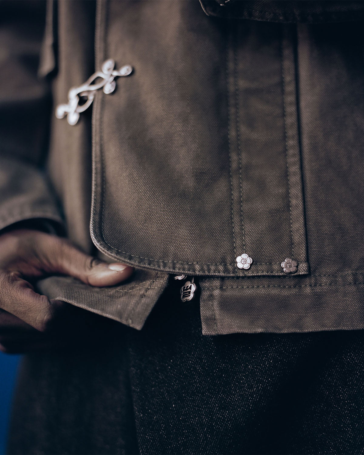 Close-up of a person wearing the Sundae School [PRE-ORDER] OLIVE FIREMAN CLASP JACKET, featuring flower-shaped metal buttons and a zipper, holding the edge of the jacket. The person pairs it with black pants.