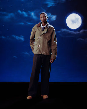 A man stands before a night sky with a full moon, wearing Sundae School's [PRE-ORDER] OLIVE FIREMAN CLASP JACKET, paired with dark wide-leg pants and brown shoes.