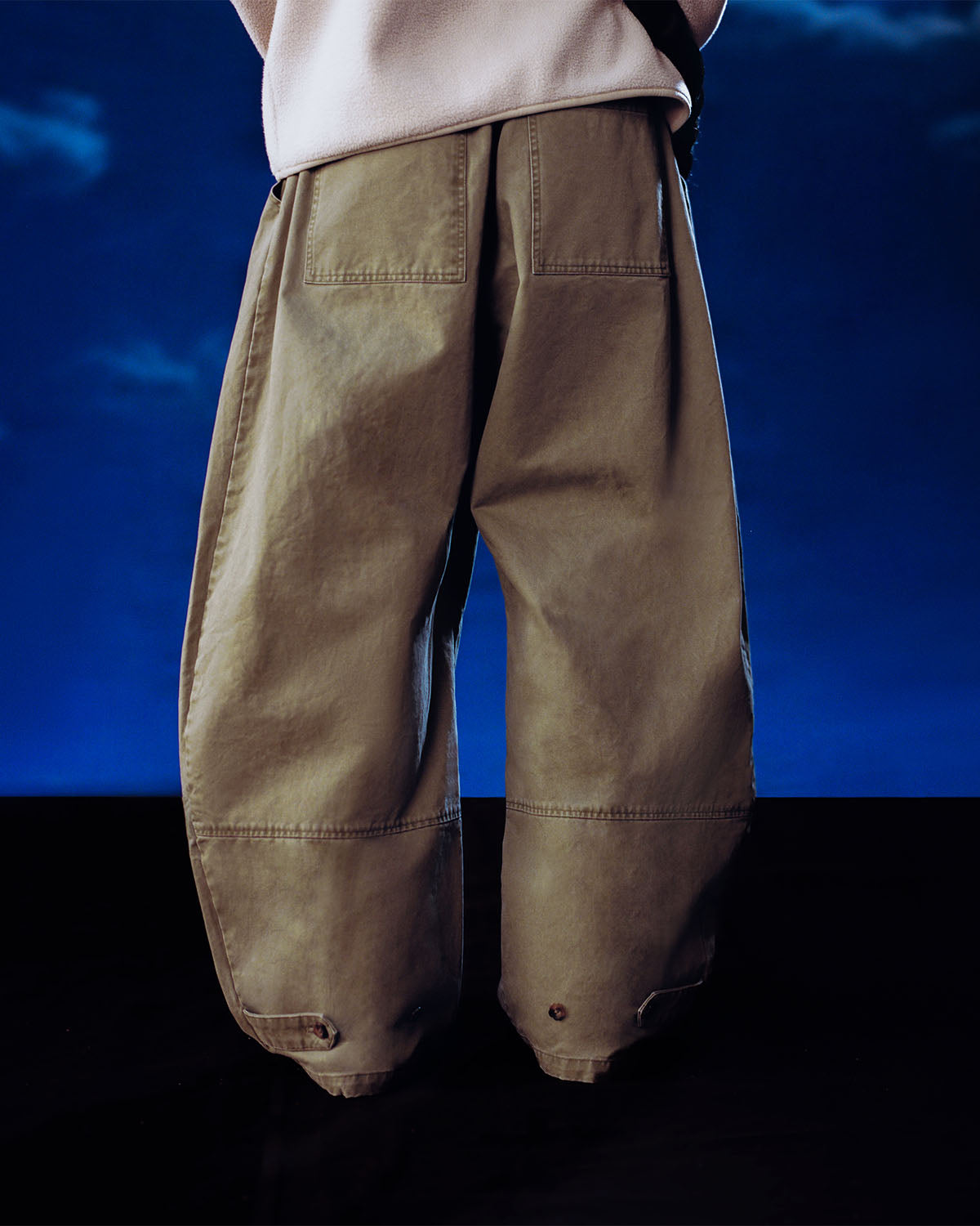 A person faces away from the camera, wearing Sundae School's OLIVE HANBOK CHINO PANTS with a hanbok-inspired silhouette and large back pockets, set against a dark blue background.