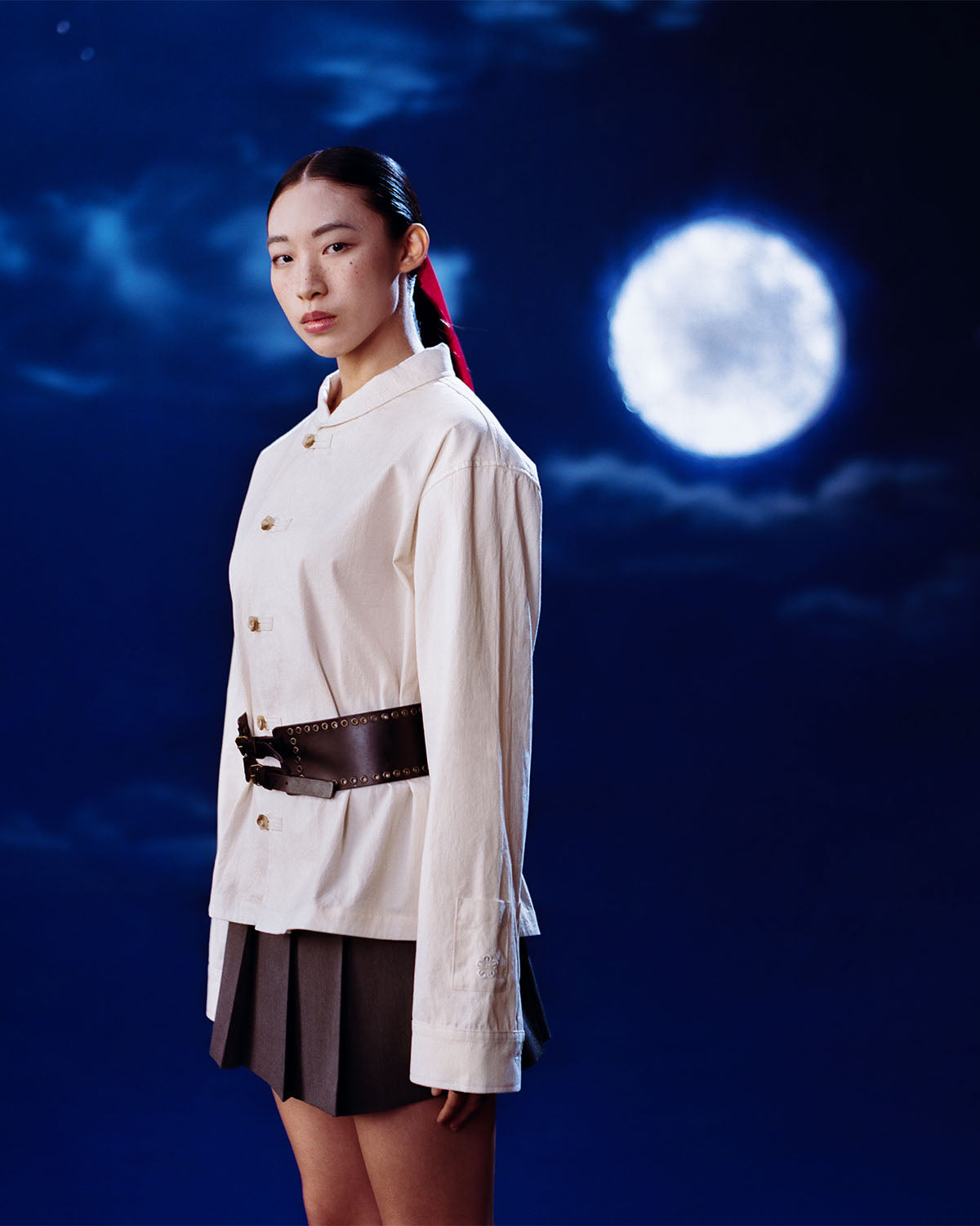 A woman with straight dark hair tied back in a red ribbon stands before a full moon, calmly gazing at the camera. She wears the Sundae School IVORY CHINA COLLAR KNOT SHACKET, styled with a black belt and pleated skirt.