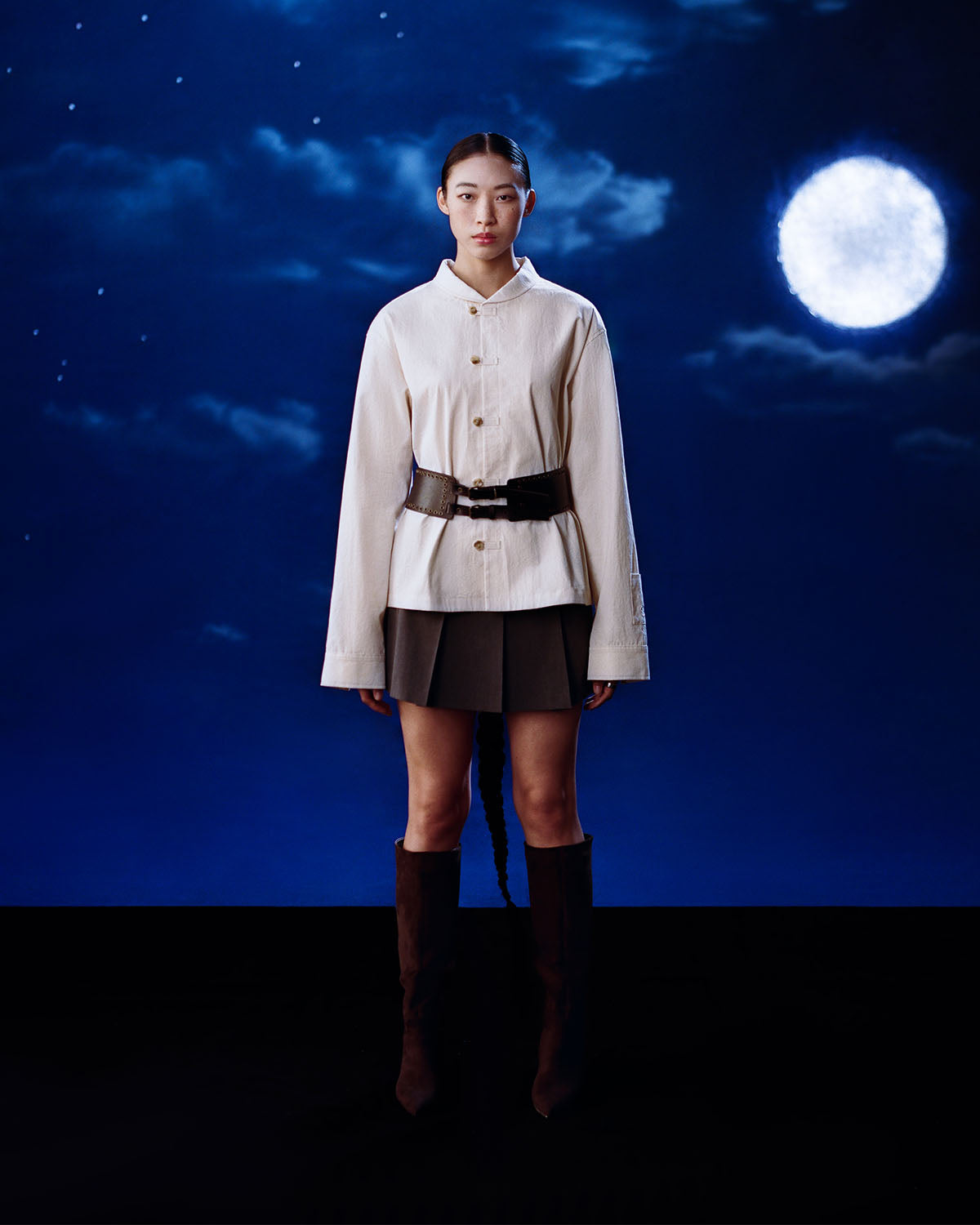 A woman stands against a dark blue night sky with clouds and a full moon, wearing the IVORY CHINA COLLAR KNOT SHACKET by Sundae School, paired with a dark pleated skirt, black belt, and tall dark boots.