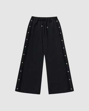 Black Fade Black Snap Sweatpants featuring vintage fade washed black cotton with custom dome snaps down both side seams, elastic waistband with drawstring ties, relaxed wide-leg fit silhouette, flat lay product shot on light background.
