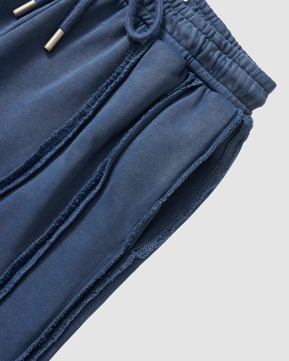 Indigo Mochi Haze Paneled Sweatpants waistband detail showing elastic band with drawstring ties and metal aglets, exposed seam construction visible, garment washed indigo cotton texture, flat lay on light background.