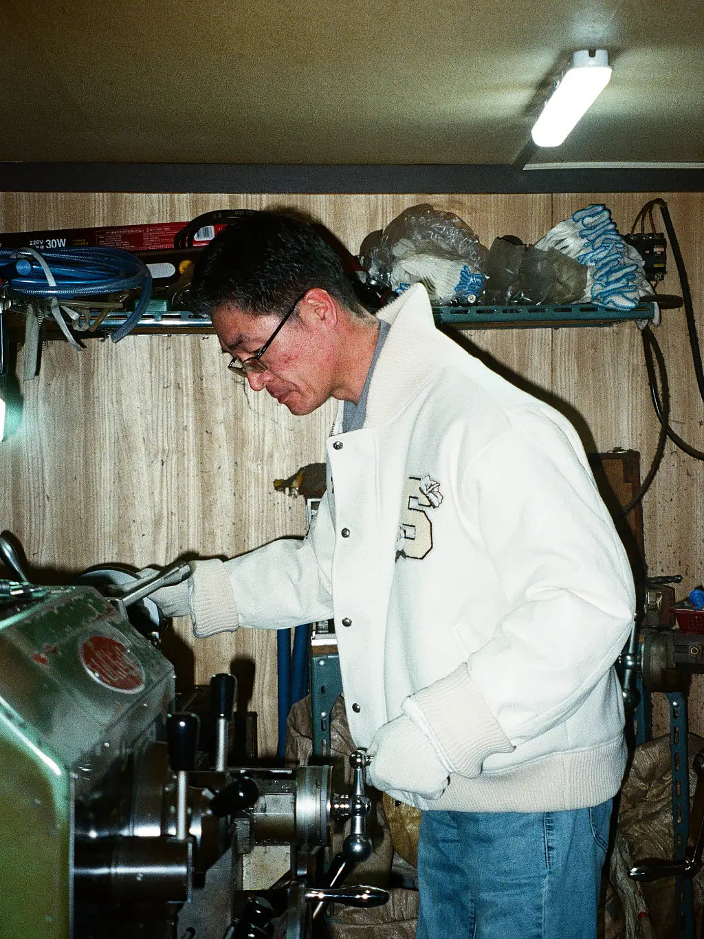 A man wearing SUNDAE SCHOOL VARSITY JACKET and blue jeans operates a metal lathe machine in a workshop with wood-paneled walls and shelves in the background.