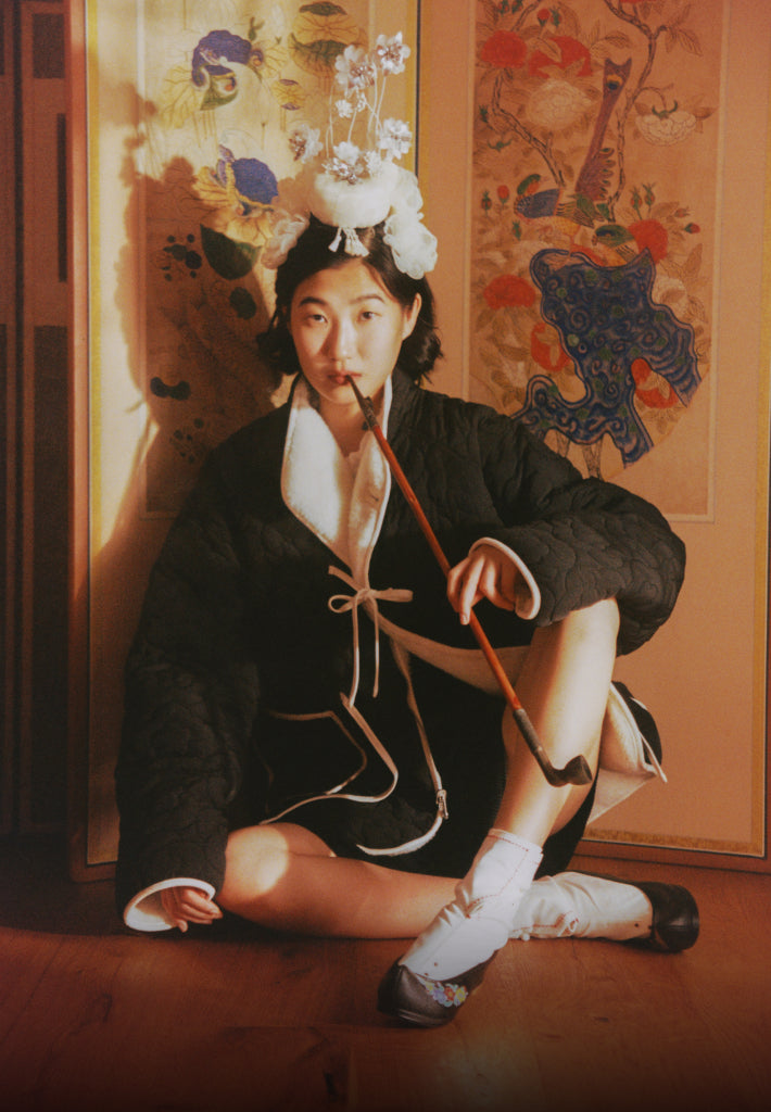 A girl wearing the black side of Black Ivory Reversible Flower Quited Jeogori Fleece biting gonbangdae, Korean traditional smoking pipe, sitting on the Korean traditional room divider.