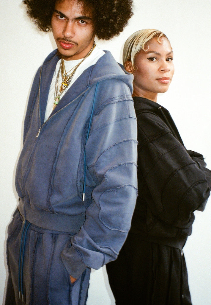 A man and a woman with their backs leaned against each other, looking at the camera, wearing Sundae School Mochi Haze Paneled Zip Up Hoodie and Mochi Haze Paneled Sweatpants in indigo blue and black. 