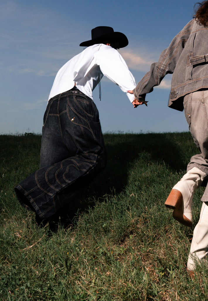A vertical mobile banner showing two people from behind holding hands as they move through a grassy field under a blue sky. The person on the left wears a black cowboy hat, a white long-sleeved shirt, and oversized, baggy dark denim jeans with decorative stitching. The person on the right is partially cropped and wears a grey denim jacket, matching pants, and off-white heeled boots.