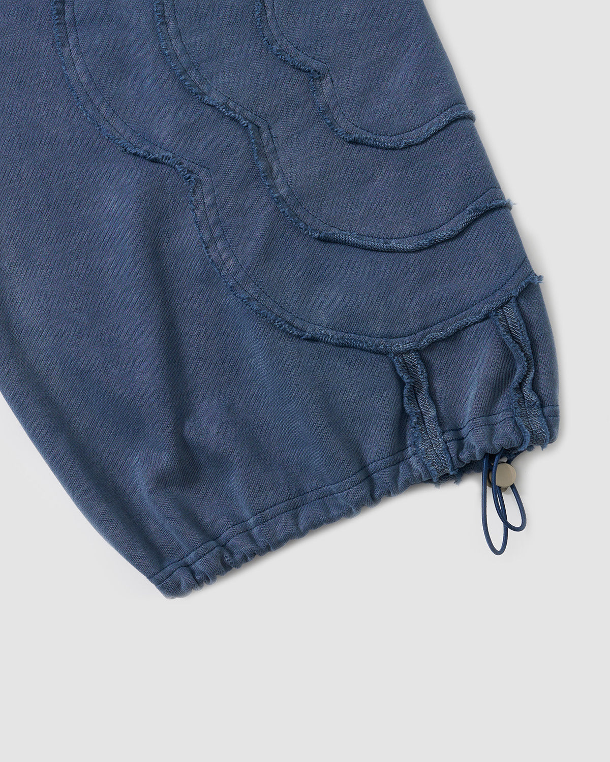 Indigo Mochi Haze Paneled Hoodie hem detail showing zip closure with branded hardware, ribbed waistband, exposed seam construction, and washed black cotton texture, flat lay on light background.