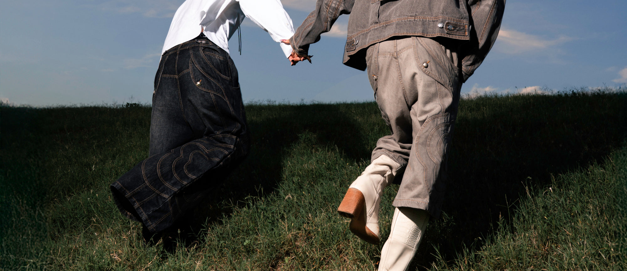 A vertical mobile banner showing two people from behind holding hands as they move through a grassy field under a blue sky. The person on the left wears a black cowboy hat, a white long-sleeved shirt, and oversized, baggy dark denim jeans with decorative stitching. The person on the right is partially cropped and wears a grey denim jacket, matching pants, and off-white heeled boots.