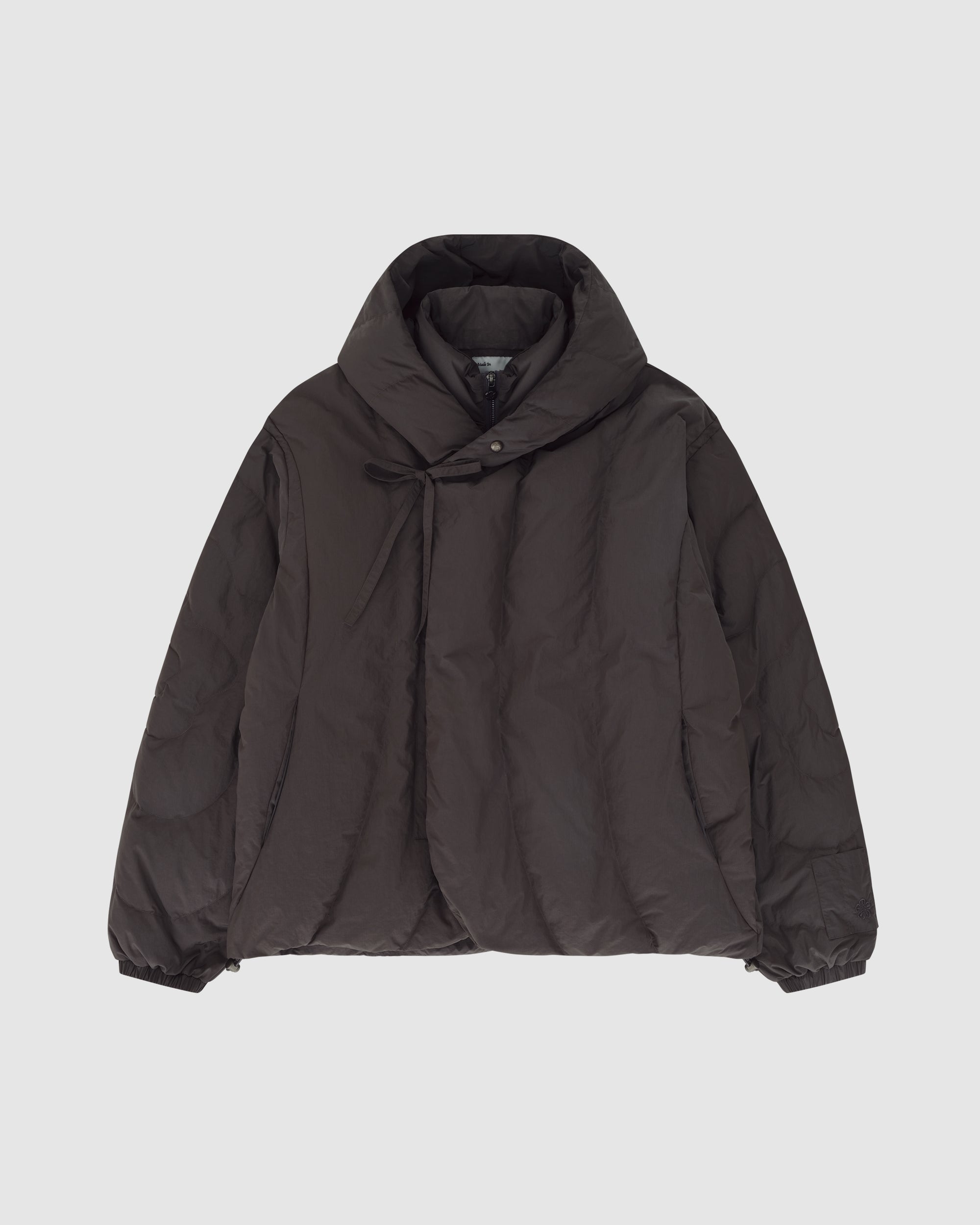 The CHARCOAL HEAVYWEIGHT MOCHI PUFFER by Sundae School, a dark brown winter jacket with a high collar, diagonal button closure, and elastic cuffs, is laid flat on a plain light gray background.