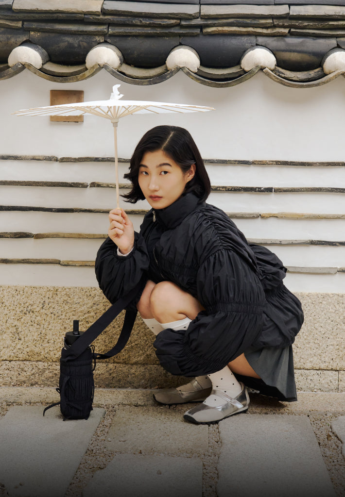 A girl squatting down in front of the Korean traditional house hanok wearing Sundae School black pleated duvet puffer, staring into the camera.