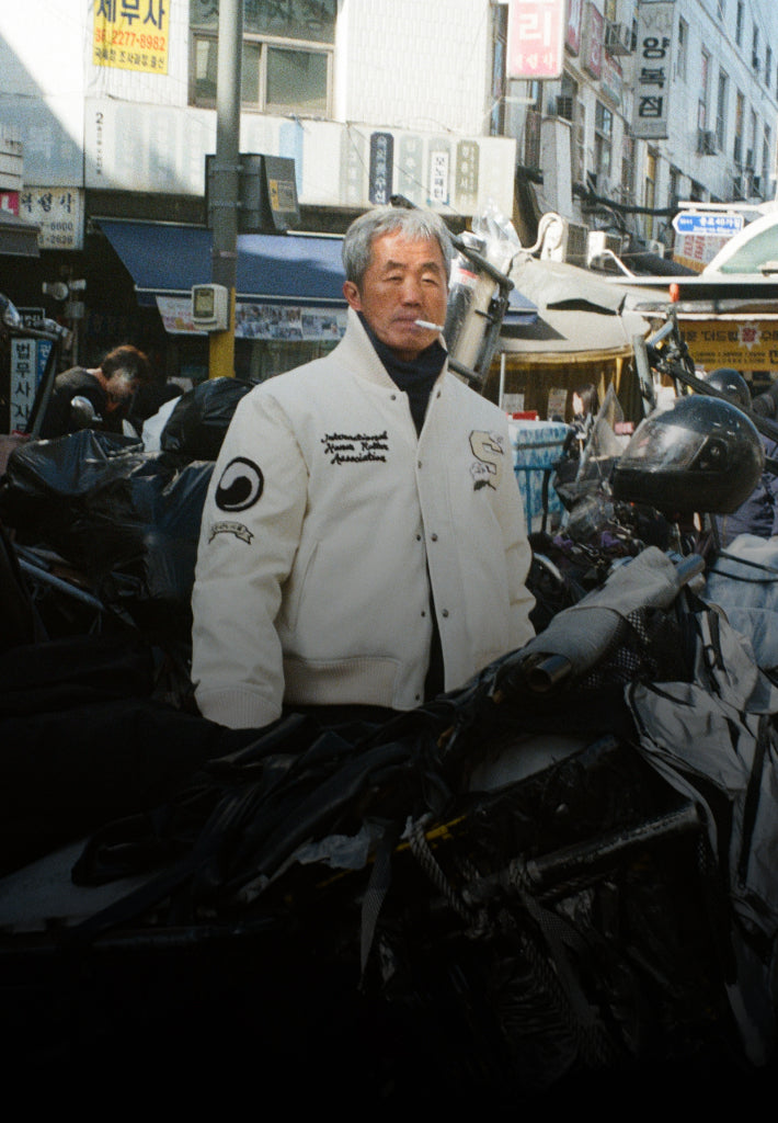 A Korean man standing behind a motorcycle with a cigarette in his mouth, wearing Sundae School Ivory Varsity Jacket in DDP, Seoul.