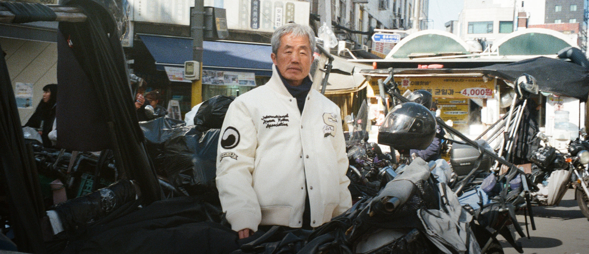 A Korean man standing behind a motorcycle staring into the camera, wearing Sundae School Ivory Varsity Jacket in DDP, Seoul.