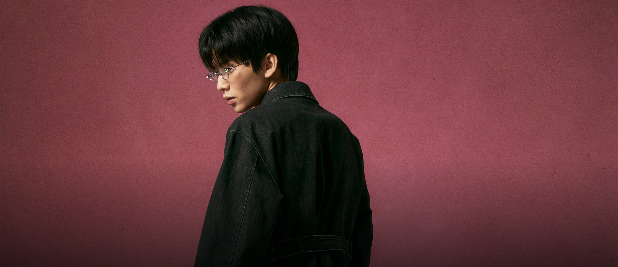 A profile shot of a model wearing a black structured work jacket and silver wire-rim glasses, looking over his shoulder against a textured pink studio backdrop.