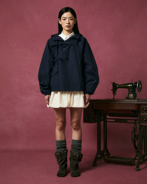 A woman poses against a mauve background wearing the Sundae School NAVY JEOGORI TIE SHELL JACKET over a cream dress, gray legwarmers, and brown boots, standing beside an antique Singer sewing machine on a wooden table.