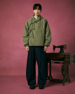 A person in a MATCHA JEOGORI TIE SHELL JACKET by Sundae School and wide-leg black pants stands on a pink floor against a pink backdrop beside an antique sewing machine on a wooden table.