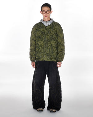 A person stands facing forward against a white background, wearing glasses, a Sundae School MATCHA MOCHI QUILTED MILITARY LINER with a floral pattern, a gray hoodie underneath, wide-leg black jeans with stitched designs, and black sneakers.