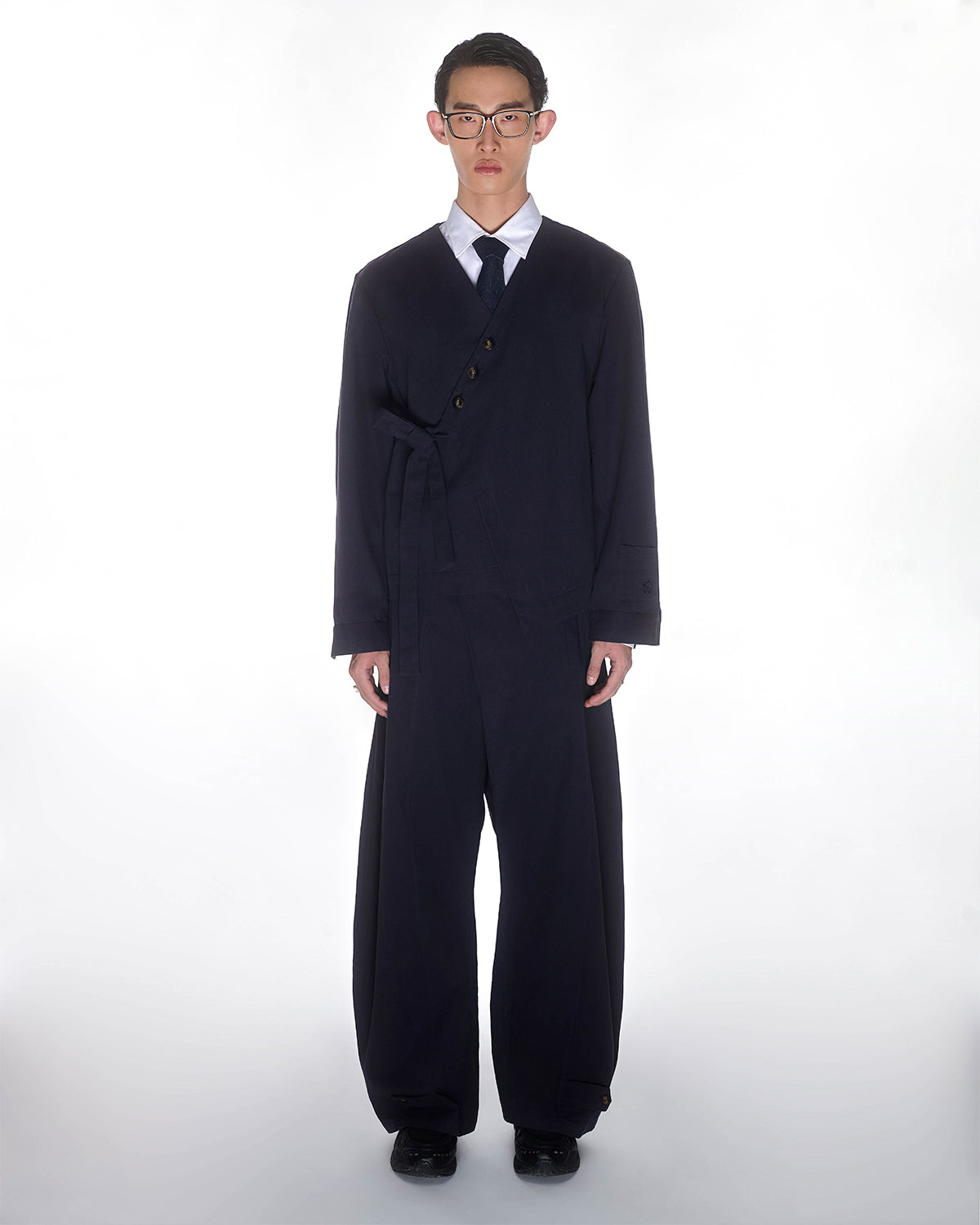 A person faces forward in a Sundae School NAVY HANBOK TIE BLAZER with an asymmetric side tie and wide-legged pants, paired with a white shirt and tie, set against a plain white background.
