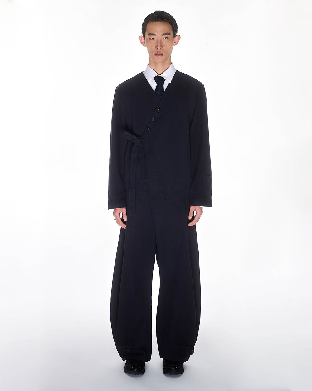 A young man stands against a white background wearing Sundae School's NAVY HANBOK BALLOON PANTS—loose-fitting, wide-leg trousers styled with a matching dark navy suit jacket that fastens asymmetrically, plus a white shirt and black tie.
