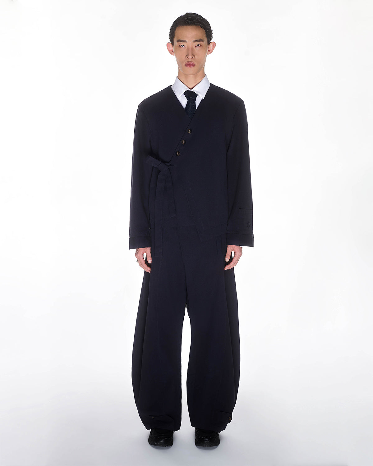 A young man stands against a white background wearing Sundae School's NAVY HANBOK BALLOON PANTS—loose-fitting, wide-leg trousers styled with a matching dark navy suit jacket that fastens asymmetrically, plus a white shirt and black tie.