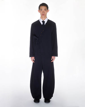 A young man stands against a white background wearing Sundae School's NAVY HANBOK BALLOON PANTS—loose-fitting, wide-leg trousers styled with a matching dark navy suit jacket that fastens asymmetrically, plus a white shirt and black tie.