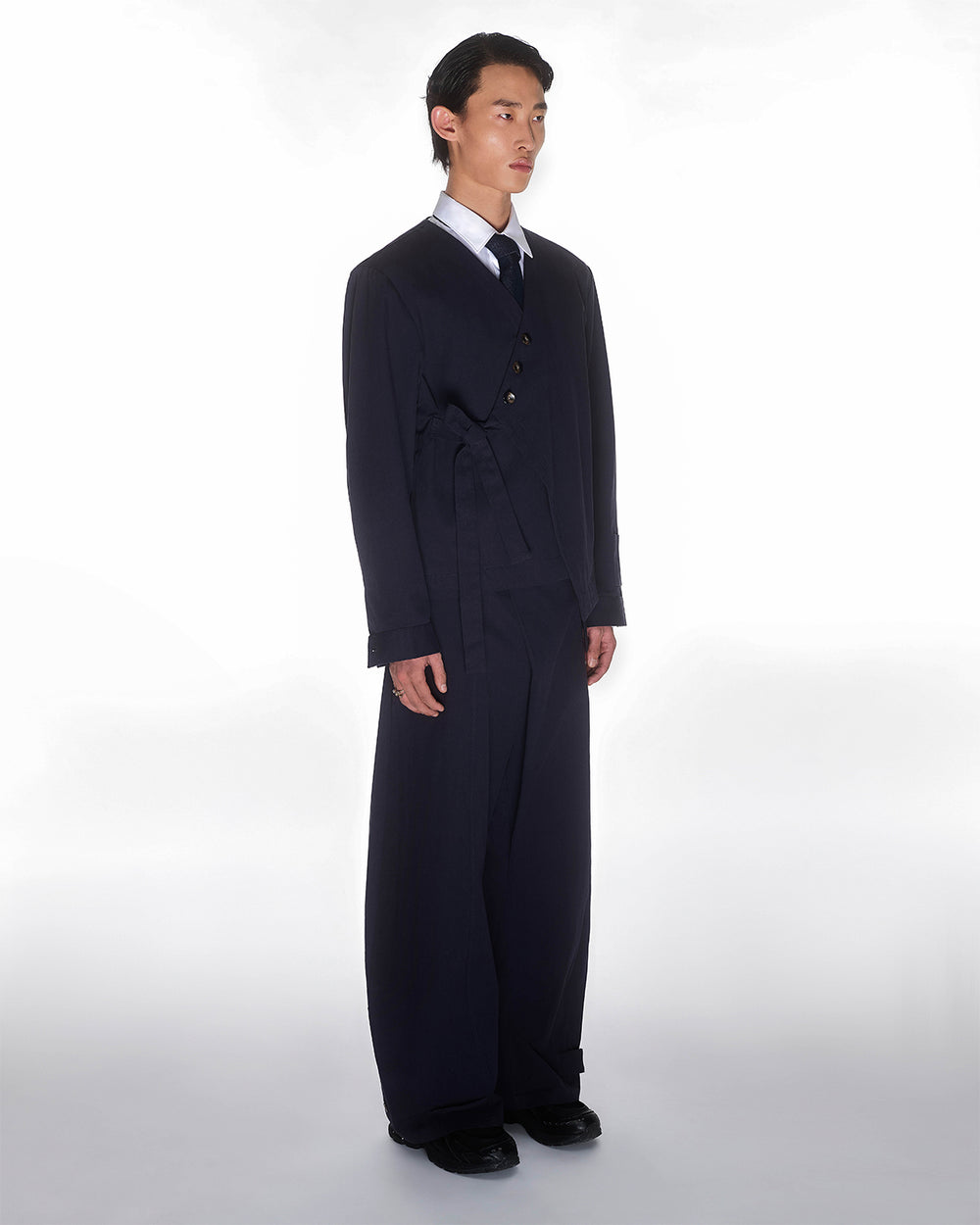 A person with short dark hair stands against a plain white background, wearing Sundae School’s NAVY HANBOK TIE BLAZER, wide-leg trousers, a white shirt, a dark tie, and black shoes.