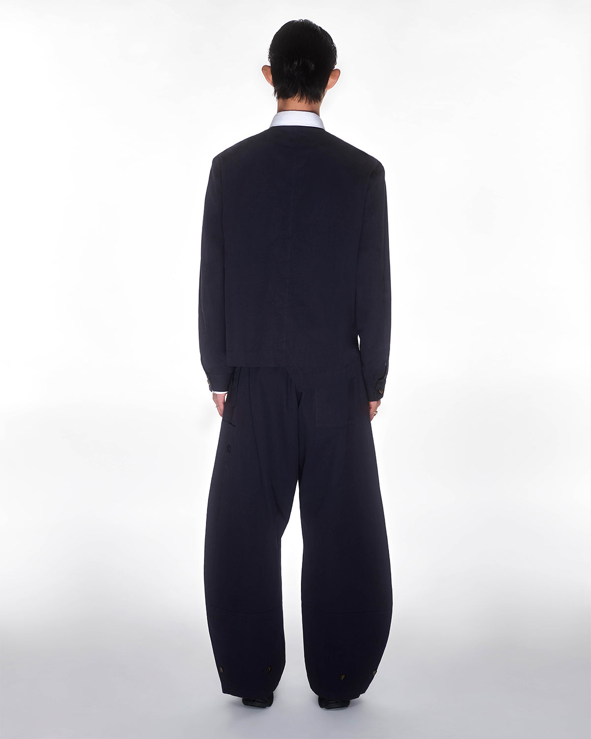 A person with dark hair, wearing a loose dark jacket and Sundae School's NAVY HANBOK BALLOON PANTS, stands facing away from the camera against a plain white background.