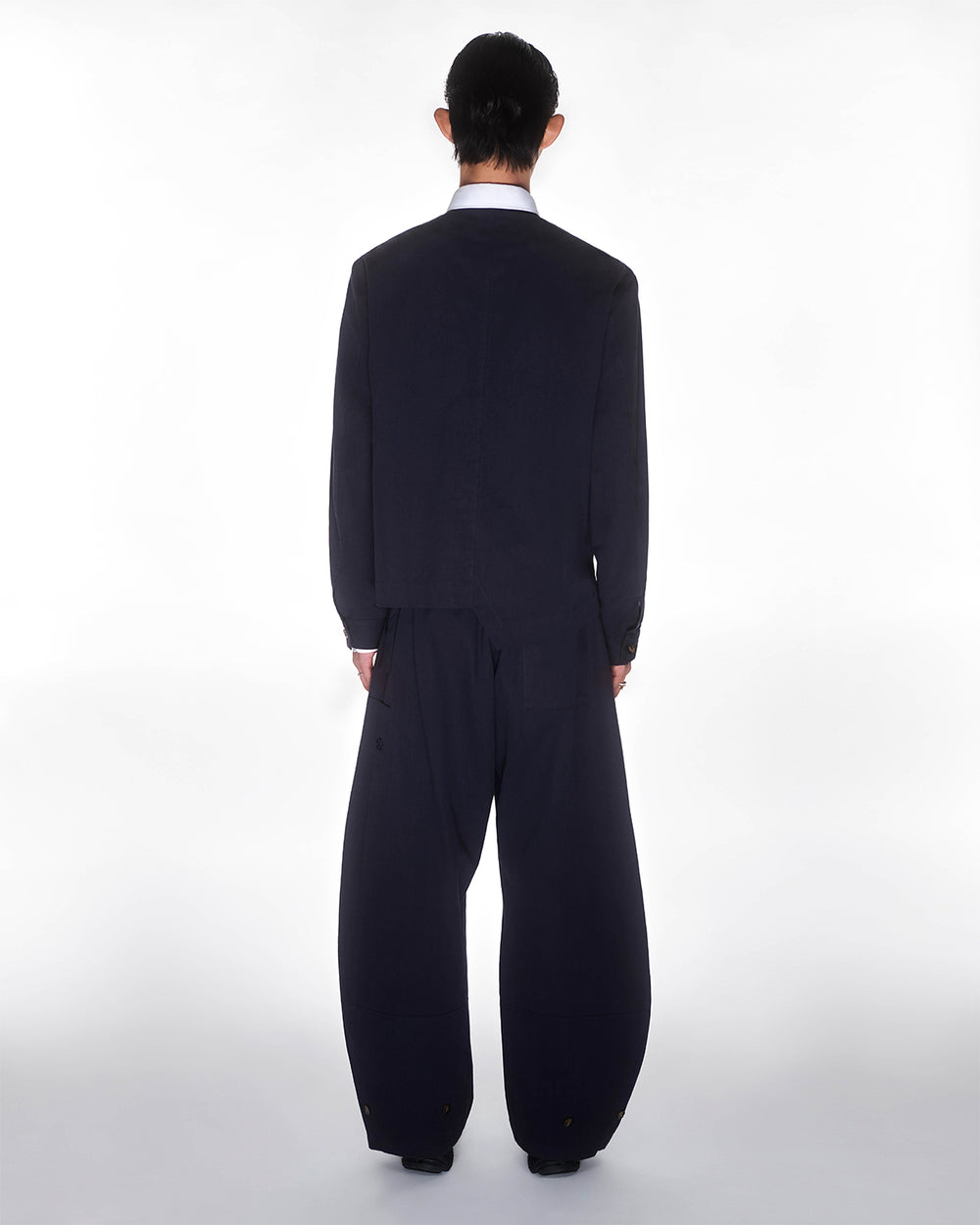 A person with dark hair, wearing a loose dark jacket and Sundae School's NAVY HANBOK BALLOON PANTS, stands facing away from the camera against a plain white background.