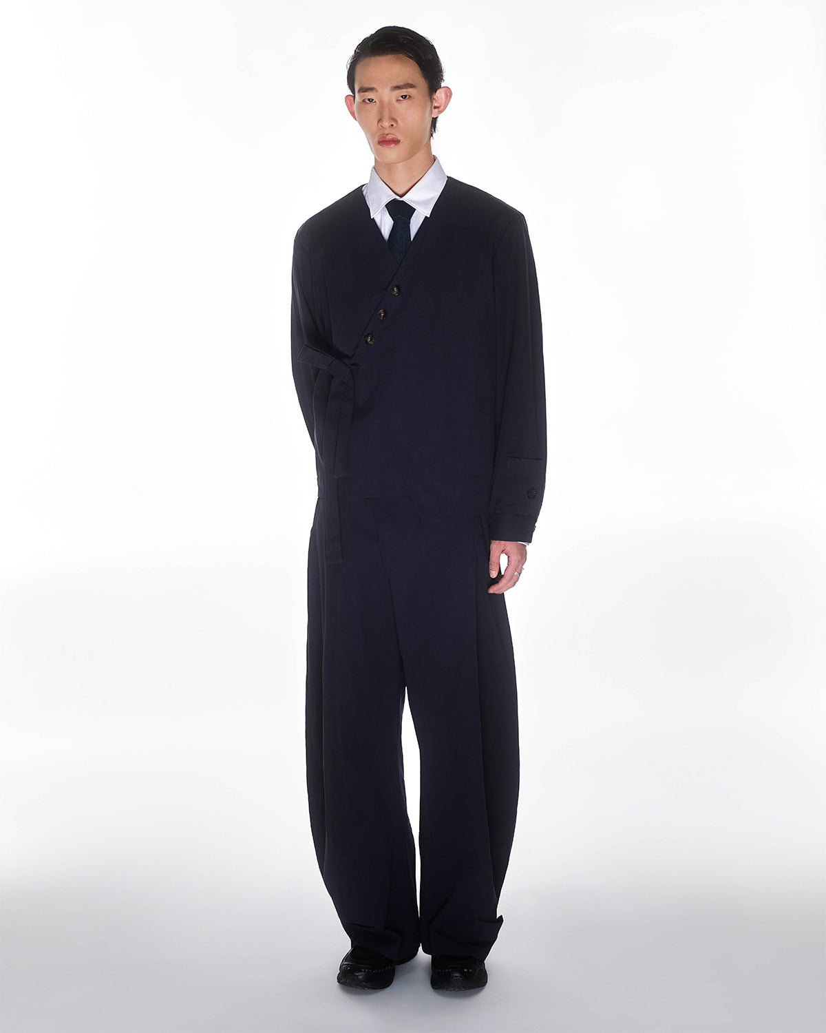 Against a white background, a person wears Sundae School's NAVY HANBOK BALLOON PANTS, paired with an oversized black suit, asymmetrical buttoned jacket, white shirt, and black tie. One arm is tucked in the jacket, and their expression is neutral.