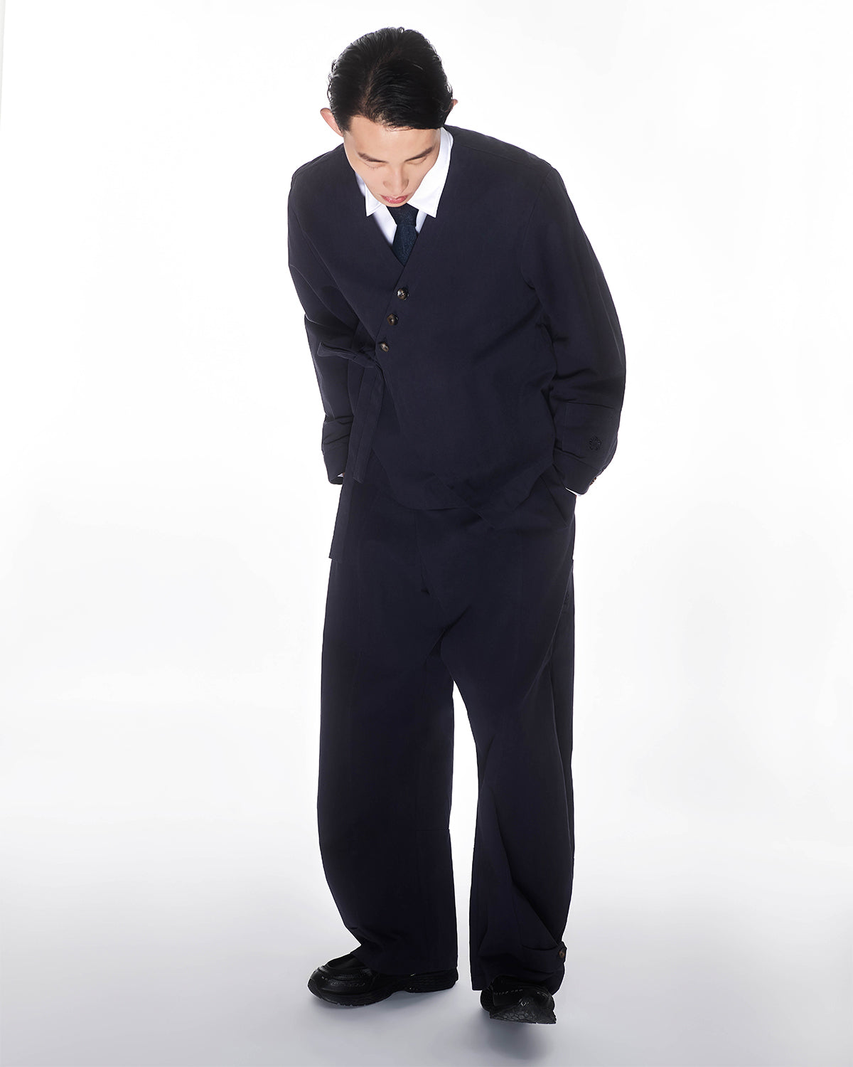 A person with dark hair wears an oversized black suit, white shirt, black tie, and Sundae School's NAVY HANBOK BALLOON PANTS, standing against a white background with hands in pockets and looking down.