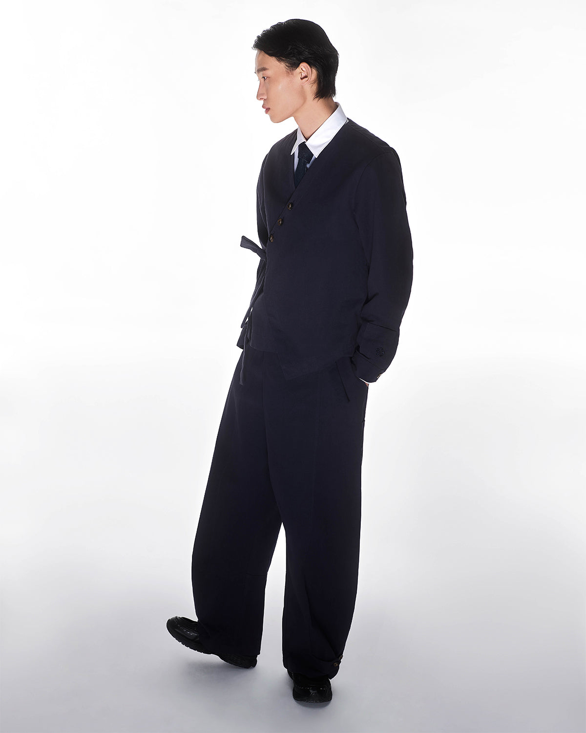 A person stands in a three-quarter pose against a plain white background, hands in pockets, wearing Sundae School's loose-fitting NAVY HANBOK BALLOON PANTS paired with a dark suit jacket, white shirt, and tie.